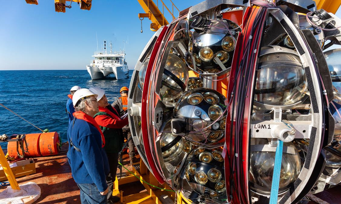 Photo prise à bord d'un bateau, on voit la mer et des scientifiques près de lignes de détection de l’expérience KM3NeT avant déploiement. Les lignes sont enroulées sur une structure sphérique qui est déposée sur le fond marin. Un robot vient alors libérer la sphère de son ancre qui en remontant par flottaison déroule la ligne de détection. Photo : Patrick Dumas/CNRS